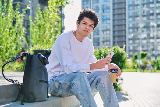 Relaxed resting handsome young male with smartphone, glass of coffee to go looking at camera, sitting on steps, outdoor. Student 19-20 years old with backpack, lifestyle, youth concept
