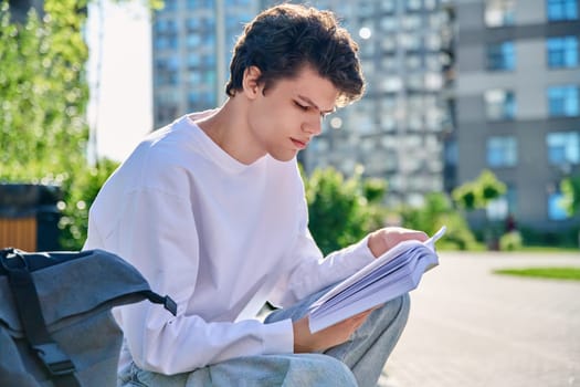 Handsome young male university college student reading a book sitting on outdoor steps. Education, knowledge, youth 19-20 years old, literary hobby and leisure