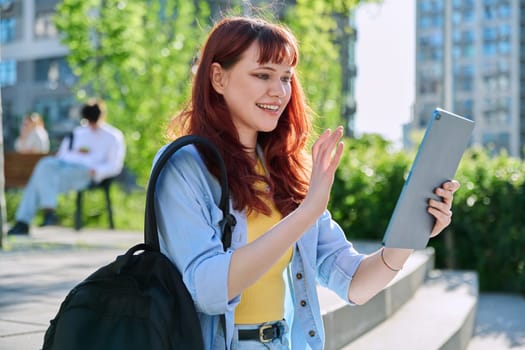 Young attractive female college student using digital tablet outdoor, talking on video call chat conference, educational building background. Education, technology, training, 19,20 years age youth concept