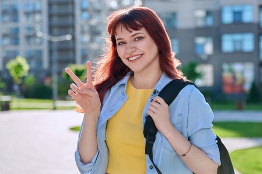Young smiling happy beautiful red-haired hipster female looking at camera showing victory peace gesture with her hand, outdoor in sunlight. Beauty, fashion, style, lifestyle youth concept