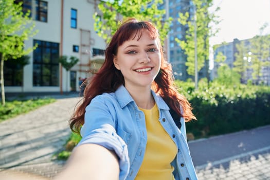 Selfie portrait of young beautiful attractive happy red-haired female looking at camera outdoor. Sunny day, teenage girl student 19-20 years old, near educational building. Education youth lifestyle