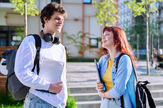 Teenage college students guy and girl talking, standing outdoor near educational building. Youth 19-20 years old, education, lifestyle, friendship concept