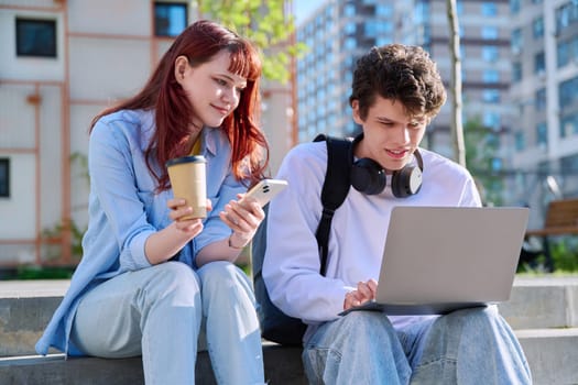 Teenage college students guy and girl talking, using laptop smartphone, sitting outdoor near educational building. Youth 19-20 years old, education, technologies, lifestyle, friendship concept