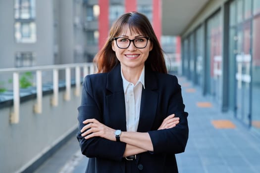 Mature confident successful business woman with crossed arms in black suit looking at camera outdoors, backdrop of modern city. Business, entrepreneurship mentoring insurance sales advertising work