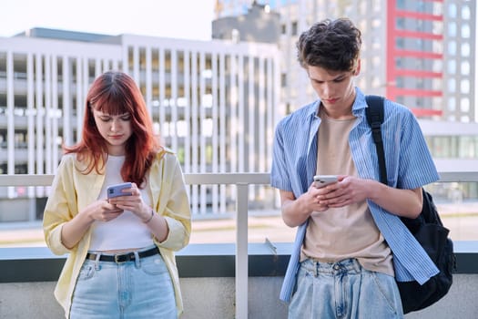Teenage friends university college students guy and girl using smartphones, urban outdoor, city modern buildings background. Technology, friendship, youth 19-20 years old, lifestyle concept