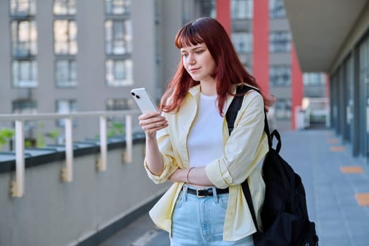 Young female using smartphone, modern city background. Teenage student 19-20 years old, texting using mobile apps applications for leisure study travel. Technology Internet education youth urban style