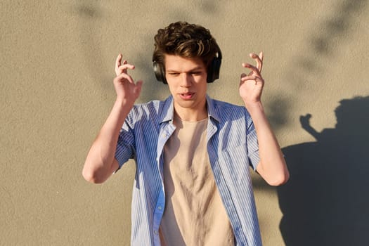 Young handsome male in wireless headphones listening to music, against the backdrop of an outdoor sunny gray wall