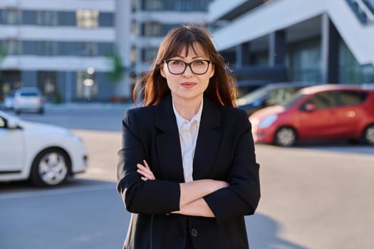 Mature confident successful business woman with crossed arms in black suit looking at camera outdoors, backdrop of modern city. Business, entrepreneurship mentoring insurance sales advertising work
