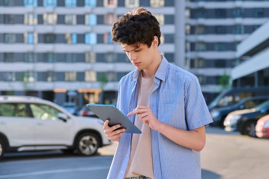 Young male using digital tablet on city street. Handsome guy student 19-20 years old, using pad for leisure, studying, working. Technology, youth, education, urban style concept