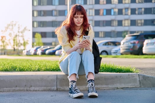 Young beautiful serious red-haired woman using smartphone sitting on floor sunny day outdoor city urban style. Youth lifestyle technology internet online applications for leisure tourism learning work