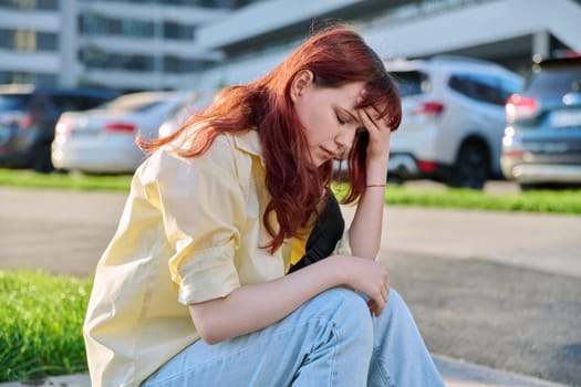 Upset sad unhappy young female sitting on steps. Pensive serious beautiful red-haired girl student 19-20 years old sitting outdoor. Problems, difficulties, depression, mental health of young people