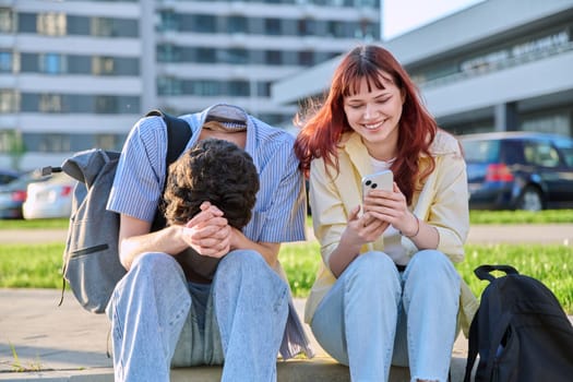 Friends young guy and girl university college students talking together, looking at smartphone outdoor, city modern buildings background. Lifestyle, communication, youth 19-20 years old, urban style