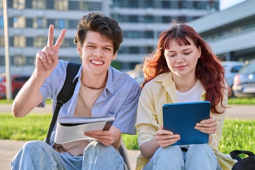 Teenage college students guy and girl talking, using digital tablet, sitting outdoor, urban modern city. Youth 19-20 years old, education, technologies, lifestyle, friendship concept