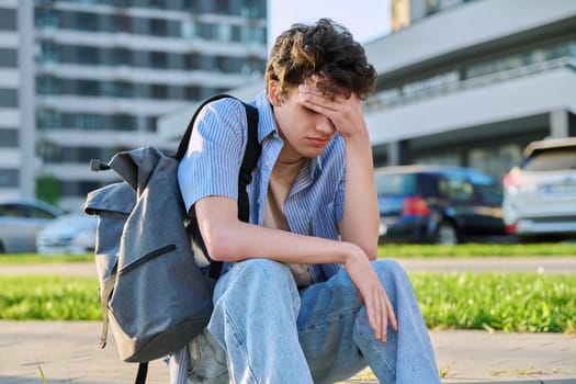 Upset sad unhappy young male sitting outdoor on steps. Guy university college student with backpack sitting, holding head with hands. Problems, difficulties, depression, mental health of young people