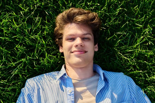 Top view, happy handsome smiling young male looking at camera, lying on background of green lawn grass.