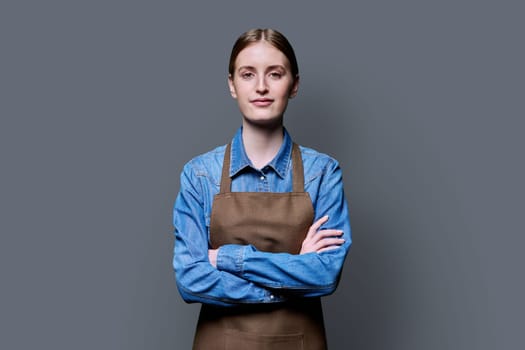 Portrait of young smiling confident woman in apron on grey studio background. Successful positive female with crossed arms. Worker, startup, small business, service sector, staff, youth concept