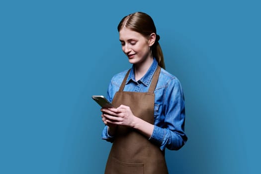 Young female worker in an apron using smartphone on blue studio background. Work, business, online internet services, mobile apps applications, technology concept