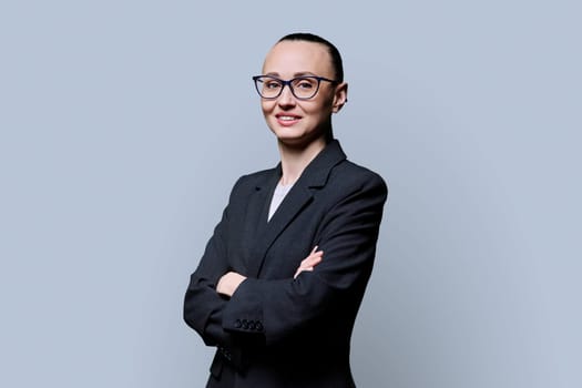 Portrait of happy smiling with teeth business woman in 30s with crossed arms, on grey studio background. Confident female in glasses, suit looking at camera. Business work job career people concept