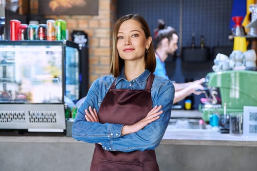 Confident successful young woman service worker in apron with crossed arms looking at camera in restaurant cafeteria coffee pastry shop. Small business, staff, occupation, entrepreneur, owner, work