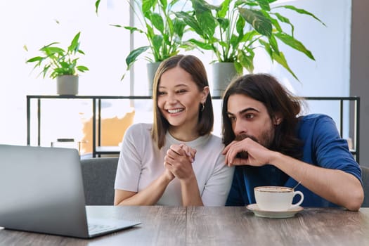 Happy smiling surprised young couple looking at laptop together while sitting in cafe, cafeteria. Leisure time for two, lifestyle, togetherness, relationship, communication, work study remotely