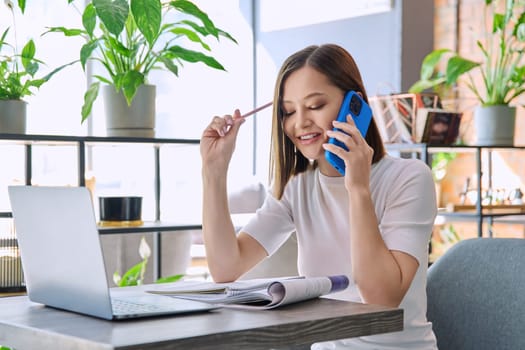 Young woman sitting at table with laptop computer talking on mobile phone in coworking cafe. Female university college student preparing for test exam, freelancer, business employee working remotely