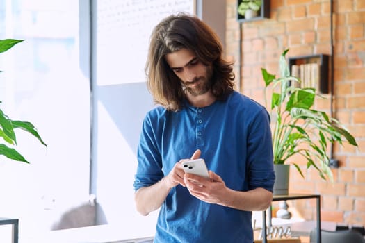 Young 30s stylish handsome serious bearded man with long hair using smartphone. Mobile technologies network Internet applications apps for leisure work business communication chatting education