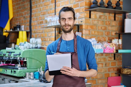 Young man in apron, food service worker, small business owner entrepreneur with work papers looking at camera near counter of coffee shop cafe cafeteria. Staff, occupation, successful business, work