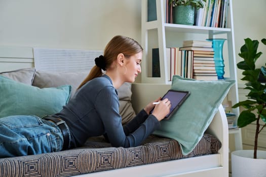 Young female teenager with digital tablet, stylus drawing illustrating lying on couch at home. Technologies, leisure, creativity, education, freelance work, lifestyle, youth concept