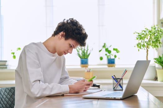 Young guy student using smartphone, sitting at his desk at home. Mobile applications apps for study education