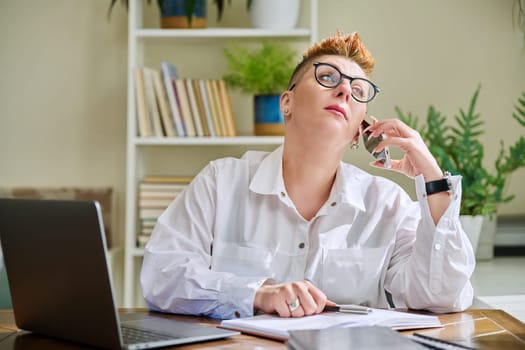 Middle-aged business woman talking on mobile phone sitting at desk with laptop computer. Serious confident mature female working with business papers. Work, workplace, 40s people