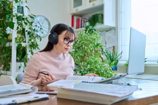 Middle aged woman teacher in headphones with textbook working online using laptop computer in home office. Video conference remote meeting consultation e lesson training education technology concept