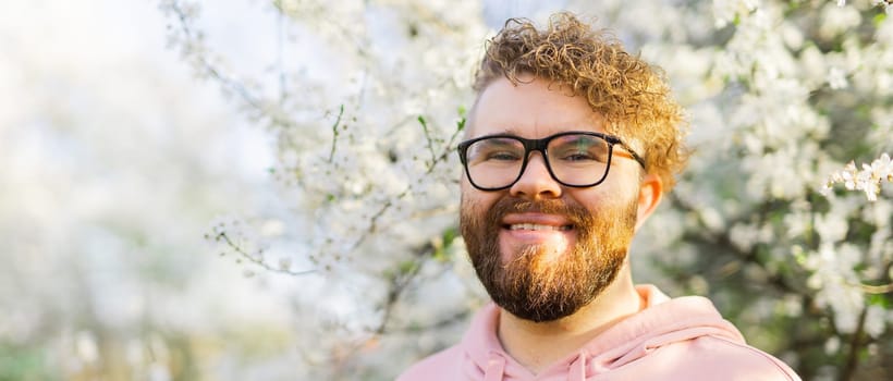 Male bearded guy standing under branches with flowers of blooming almond or cherry tree in spring garden. Spring blossom. Copy space