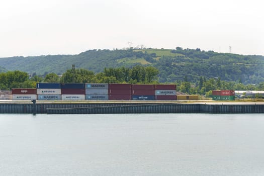Lyon, France - May 30, 2023: Stacked shipping containers in varying colors beside a calm river, with a green hill in the background under a clear sky.