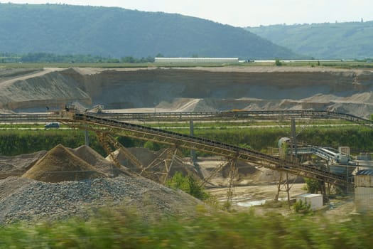 Conveyor belts transport materials in an open-pit mining operation under clear skies, with hills in the background.