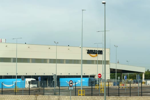 Montelimar, France - May 30, 2023: A broad view of a large Amazon warehouse with its iconic logo, surrounded by security fences and street lamps against a cloudy sky.