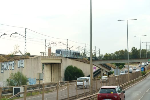 Seville, Spain - June 1, 2023: A train travels across an overpass above a road lined with vehicles, showcasing daily transportation dynamics.