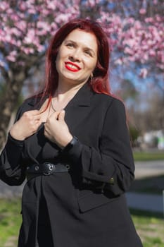 Fashion outdoor photo of beautiful woman with red curly hair in elegant suit posing in spring flowering park with blooming cherry tree. Copy space and empty place for advertising text.