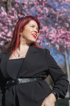 Fashion outdoor photo of beautiful woman with red curly hair in elegant suit posing in spring flowering park with blooming cherry tree. Copy space and empty place for advertising text.