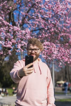 Spring day. Bearded man in pink shirt talking by phone. Spring pink sakura blossom. Handsome young man with smartphone. Fashionable man in trendy glasses. Bearded stylish man. Male fashion.