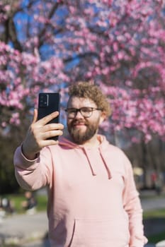 Happy curly man takes selfie against backdrop of flowering tree in spring for his internet communications. Weekend and social networks