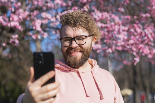 Happy curly man takes selfie against backdrop of flowering tree in spring for his internet communications. Weekend and social networks