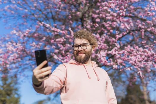 Spring day. Bearded man in pink shirt talking by phone. Spring pink sakura blossom. Handsome young man with smartphone. Fashionable man in trendy glasses. Bearded stylish man. Male fashion.