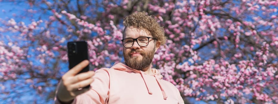 Spring day. Bearded man in pink shirt talking by phone. Spring pink sakura blossom. Handsome young man with smartphone. Fashionable man in trendy glasses. Bearded stylish man. Male fashion.