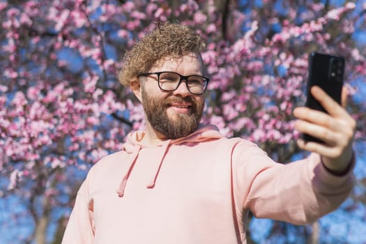 Spring day. Bearded man in pink shirt talking by phone. Spring pink sakura blossom. Handsome young man with smartphone. Fashionable man in trendy glasses. Bearded stylish man. Male fashion.