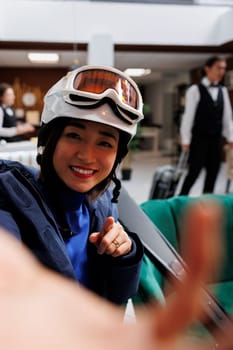 Youthful asian woman at exclusive ski resort holding cellphone for selfie picture. Enthuisiastic female traveler wearing winter jacket ski goggles and helmet ready for wintersport activities.