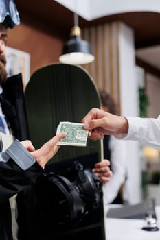 Close-up of male traveler with snowboard giving tip money to employee at counter of luxury winter resort. Detailed shot showing man wearing snowboarding gear tipping staff with cash in hotel lobby.