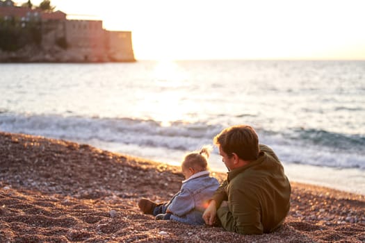 Dad is lying on the beach next to a little girl sorting through the pebbles. Back view. High quality photo