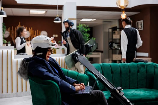 Couch in lounge area provides comfort for asian woman in snow gear as she uses her laptop. Female traveler searches for winter activities at ski resort through minicomputer with skiing skis nearby.