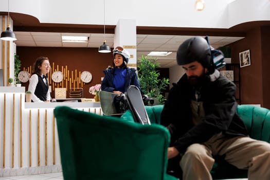 In lounge area friendly concierge assists female tourist with check in at ski resort while young man adjusts winter gear on sofa. Two travellers in hotel reception ready for fun wintersports.