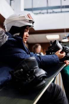 Winter-ready female traveler adjusting and ensures safety of snowboard boots in hotel lobby. Youthful woman maintaining snowboarding equipment, anticipating wintersports adventure.